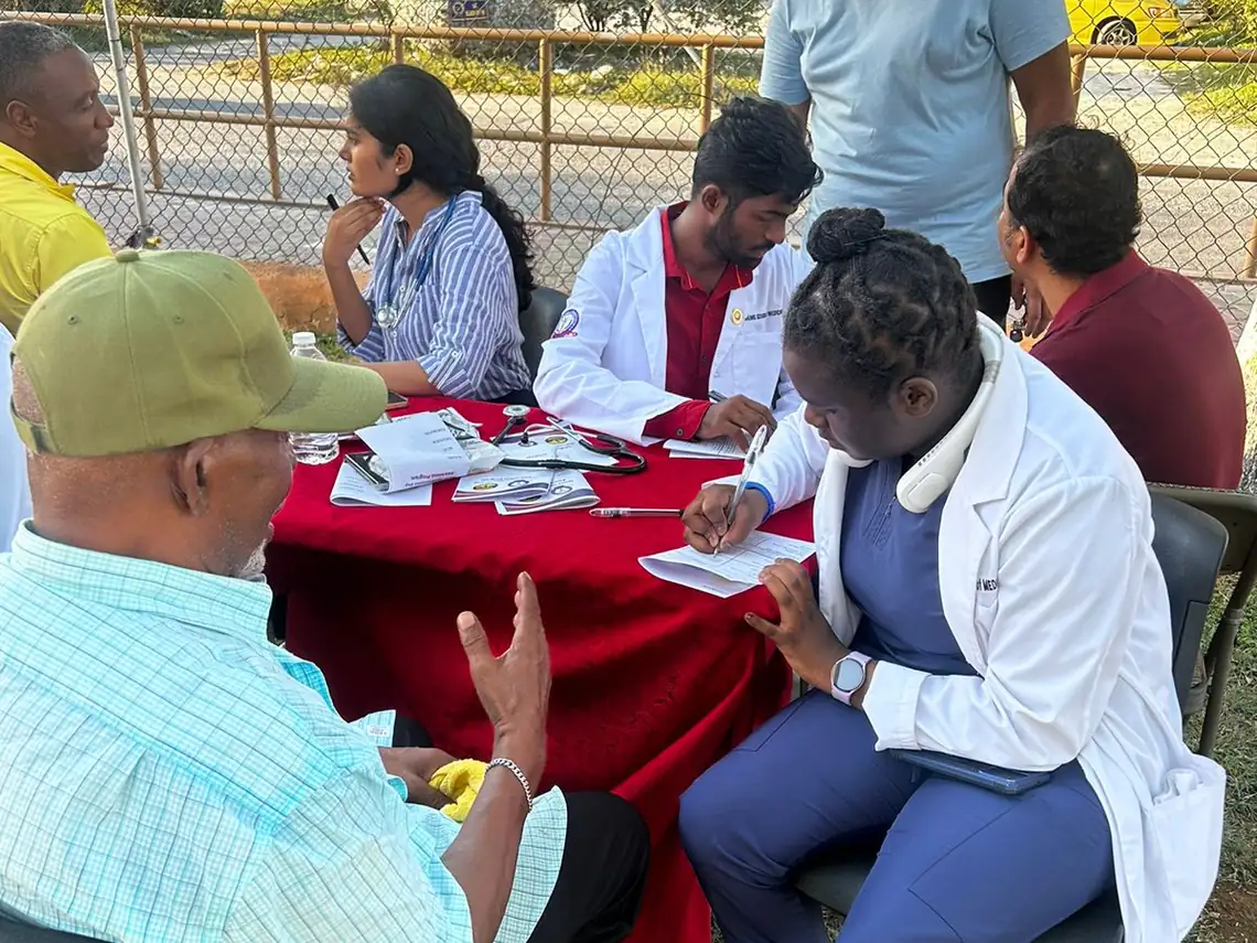 An AAIMS student in a white coat taking notes while an older resident speaks to her at a health-fair table.